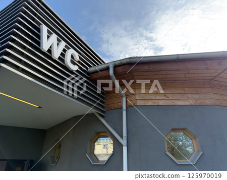 Exterior view of public restroom with large WC sign, modern horizontal paneling, wooden roof trim and octagonal windows, representing clean architectural solution for functional city service building 125970019