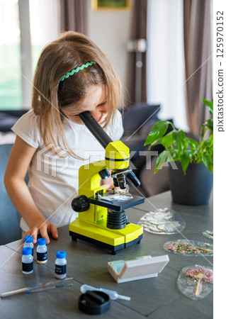 Little girl examining plant samples under a microscope at home. Concept of early science education and botanical exploration Little girl examining plant samples under a microscope at home. Concept of early science education and botanical exploration 125970152