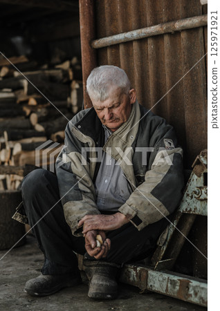 A pensioner rests on the ground, his gaze lost in thought. The elderly man wears clothes that have seen many years of work. 125971921