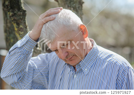 A portrait of a pensioner stroking his gray hair. A sad old man standing by a tree. A portrait of a pensioner stroking his gray hair. A sad old man standing by a tree. 125971927
