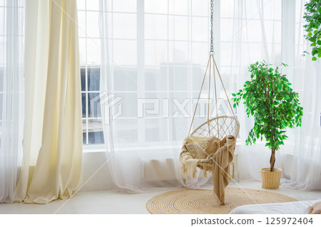 Cozy hanging wicker chair in living room with natural light, greenery on big window background 125972404