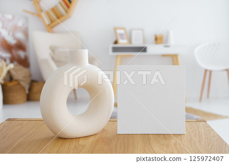 Decorative vase and blank white cardboard on wooden table in bright living room. Mockup, template 125972407