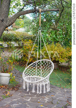 Comfortable hanging wicker white chair, hammock chair in boho style hung on a tree in summer garden Comfortable hanging wicker white chair, hammock chair in boho style hung on a tree in summer garden 125972410