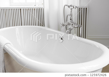 Shiny metal chrome faucet, elegant water tap on white freestanding bathtub in bathroom 125972426