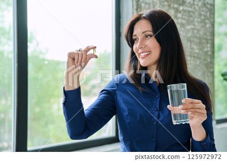 Beautiful woman taking omega 3 capsule fish oil pill, looking at camera with glass of water 125972972