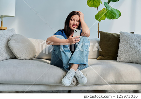 Young confused woman sitting at home on the couch using smartphone 125973010