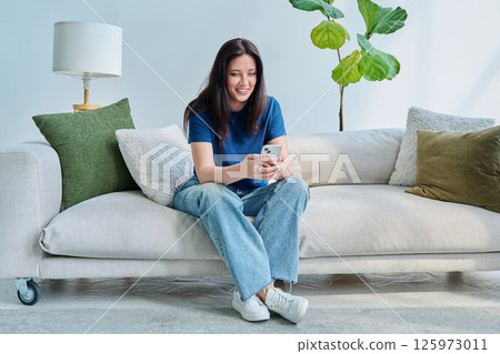 Young happy woman sitting at home on the couch using a smartphone 125973011