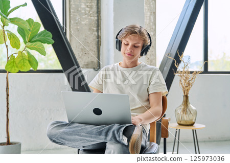 Handsome smiling teenage boy in headphones sitting at home typing on laptop Handsome smiling teenage boy in headphones sitting at home typing on laptop 125973036