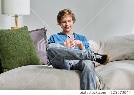 Handsome smiling teenage guy using smartphone sitting on couch at home 125973044