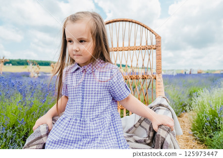 A little girl sits in a rocking chair in a lavender field. A girl with long hair and a beautiful purple dress is resting near a field covered with purple flowers. 125973447