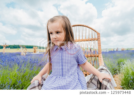 A little girl sits in a rocking chair in a lavender field. A girl with long hair and a beautiful purple dress is resting near a field covered with purple flowers. 125973448