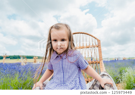 A little girl sits in a rocking chair in a lavender field. A girl with long hair and a beautiful purple dress is resting near a field covered with purple flowers. 125973449
