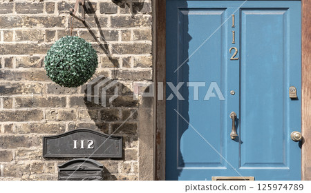 Blue composite door is closed with Door knocker, Letter box and Artificial topiary ball decoration on Brick wall front of the House number 112. Blue composite door is closed with Door knocker, Letter box and Artificial topiary ball decoration on Brick wall front of the House number 112. 125974789