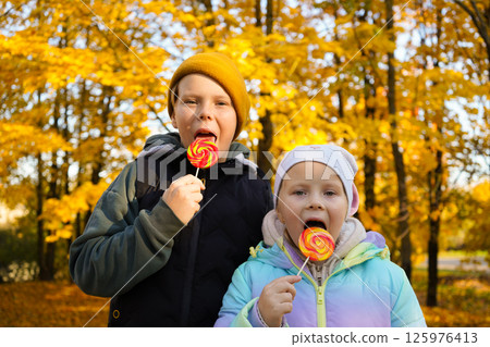 Portrait of a boy and a girl eating lollipops in an autumn park. Portrait of a boy and a girl eating lollipops in an autumn park. 125976413