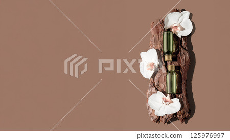 Two green glass cosmetic bottles on textured tree bark podium with white orchid flowers in rays of sunlight with hard shadows. 125976997