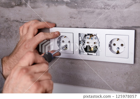 A man uses a screwdriver to install an electrical outlet on a porcelain tile wall 125978057