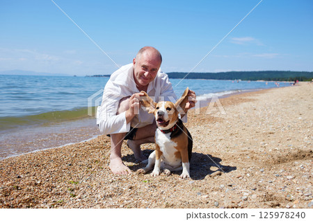 A happy man enjoys a sunny day on the sea beach with his beagle dog on a warm summer day 125978240