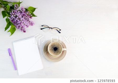 Lilac branch, cup of coffee, a glasses, notebook with pen on a white wooden background. Spring background. 125978593