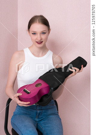 Attractive young female musician is taking ukulele out of black soft case and looking at camera. Cheerful young adult woman musician in casual clothing with white tank top and blue jeans 125978705