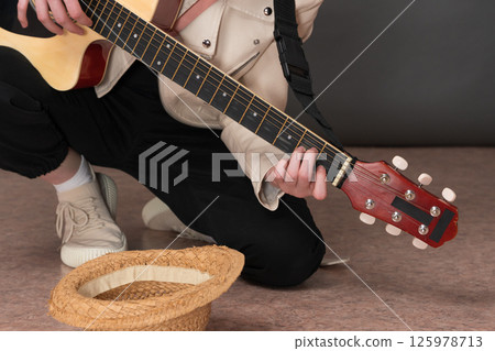 Female guitarist street musician playing acoustic guitar for money and in front of her lying empty straw hat for donation for playing guitar. Unrecognizable woman kneeling while plucking instrument 125978713