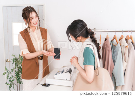 一位女店員在收銀台用智慧型手機向一位女顧客解釋某件事 一位女店員在收銀台用智慧型手機向一位女顧客解釋某件事 125979615