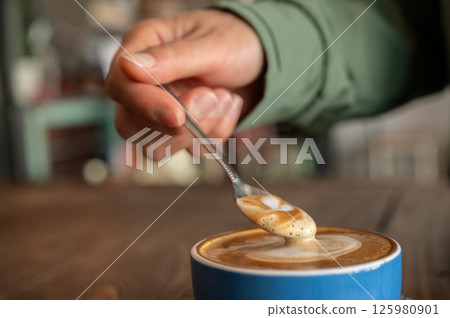 Close up of someone scooping creamy surface of hot latte coffee. A latte is a coffee drink made with espresso and steamed milk. 125980901