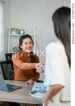 Two women engage in a professional handshake during a business meeting in a contemporary office environment. 125981929
