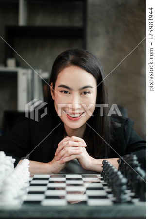 A woman sits confidently at a chessboard, ready to make her move in a modern office environment. A woman sits confidently at a chessboard, ready to make her move in a modern office environment. 125981944