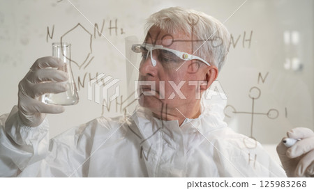 An elderly Caucasian male chemist in a protective suit looks at a test tube and writes formulas on a glass wall.  125983268