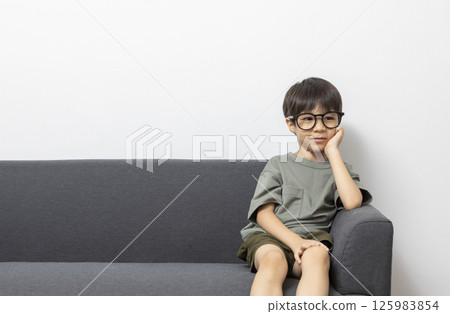 Cute Asian boy with eyeglasses sitting on a sofa at home. 125983854