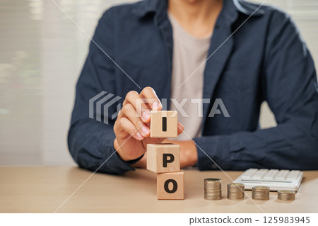 Close up businessman hand holding wooden cube blocks spelling IPO near coins and a calculator. Represents Initial Public Offering, planning, employee initiatives, and business financial accountability 125983945