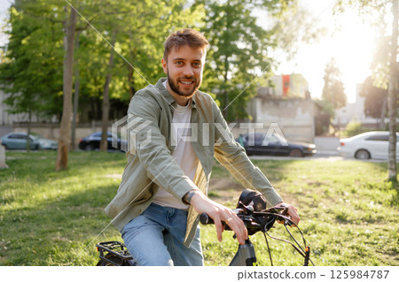 Young man enjoying a sunny day while biking in a park near urban surroundings and tall trees 125984787