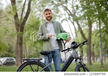 Smiling man holds green helmet next to electric bike in a sunny park with lush trees Smiling man holds green helmet next to electric bike in a sunny park with lush trees 125984799