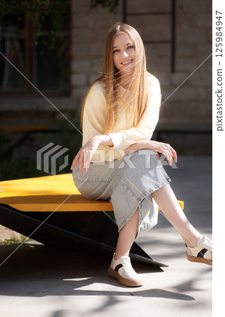 Young woman sitting on modern bench in urban park enjoying sunny day outdoors 125984947