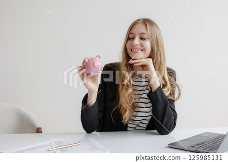 Woman holding piggy bank in modern office while smiling and looking satisfied with financial planning Woman holding piggy bank in modern office while smiling and looking satisfied with financial planning 125985131