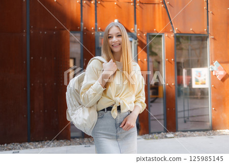 Young woman smiles while standing in front of modern architecture during the day with a backpack Young woman smiles while standing in front of modern architecture during the day with a backpack 125985145