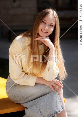 Smiling young woman sitting outdoors on a sunny day in a casual outfit enjoying the moment 125985150