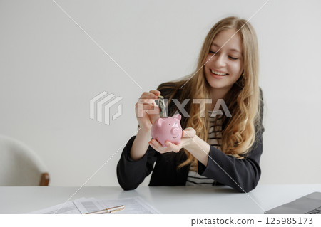Young woman saving money in a pink piggy bank at a desk with financial documents 125985173