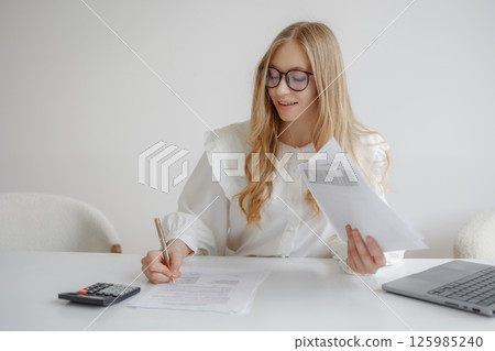 Young woman reviewing documents and writing notes in a bright home office setting with a laptop and calculator 125985240