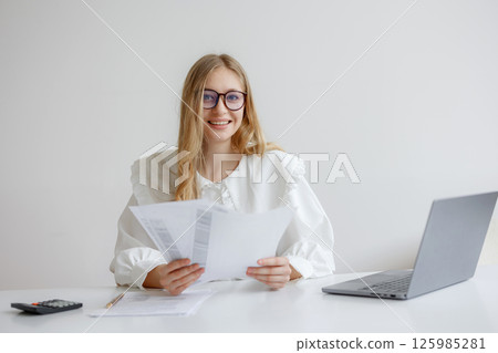 Woman reviewing documents at a desk with a laptop in a bright office environment during the day while smiling Woman reviewing documents at a desk with a laptop in a bright office environment during the day while smiling 125985281