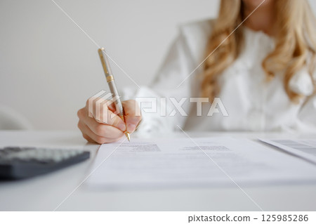 Individual completing paperwork with pen while sitting at desk with calculator and documents in bright workspace 125985286