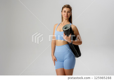 Woman holding yoga mat in light studio showcasing fitness attire and healthy lifestyle choices 125985288