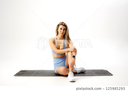 Fitness enthusiast in light blue athletic wear sitting on a mat in a bright indoor studio during a workout session Fitness enthusiast in light blue athletic wear sitting on a mat in a bright indoor studio during a workout session 125985293