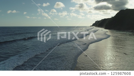 Gentle waves rolling towards the shore of Bali, Indonesia at sunset, creating a beautiful seascape with cliffs in the background, under a cloudy sky 125987608