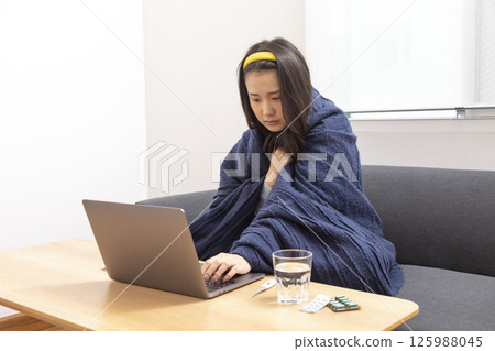 girl is sitting at sofa and working remotely on laptop. Female is blowing out snot, having fever and headache. Young woman is treated at home, took sick leave. Winter cold and flu concept girl is sitting at sofa and working remotely on laptop. Female is blowing out snot, having fever and headache. Young woman is treated at home, took sick leave. Winter cold and flu concept 125988045