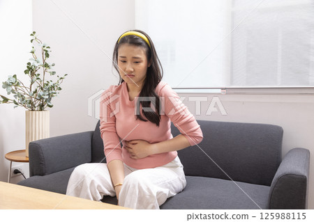 Asian attractive girl with painful face, clasping belly feel suffer from severe abdominal pain. Stressed young woman sit on sofa at home having stomach ache or periods. Menstrual gynecological concept Asian attractive girl with painful face, clasping belly feel suffer from severe abdominal pain. Stressed young woman sit on sofa at home having stomach ache or periods. Menstrual gynecological concept 125988115