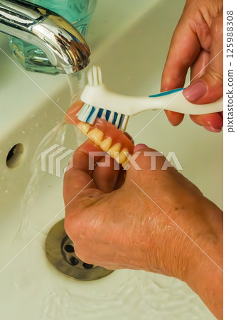 Hand is brushing dentures under running water at a bathroom sink with faucet. High quality photo 125988308