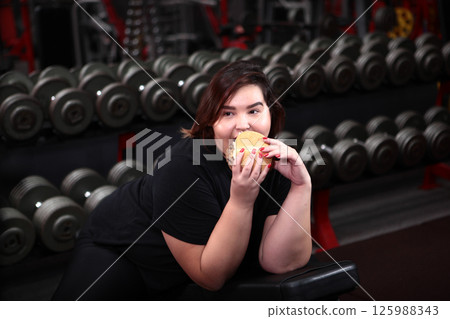 Fat, cheerful young woman of large size eats hamburger and does sports. Problem of being overweight. Concept of body positive. Fat, cheerful young woman of large size eats hamburger and does sports. Problem of being overweight. Concept of body positive. 125988343