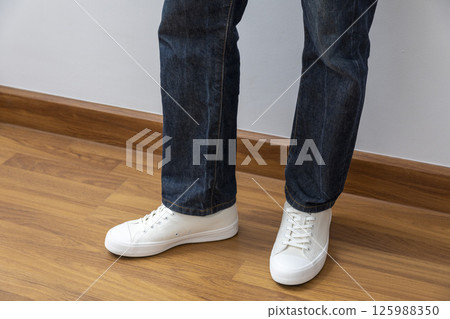 Feet wearing white chunky sole sneakers and blue jeans on the white wood floor Feet wearing white chunky sole sneakers and blue jeans on the white wood floor 125988350