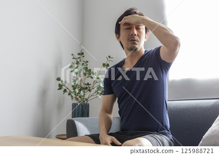 Upset sick stressed young caucasian man in casual clothes sits on sofa in the living room, bending over, massaging his head, suffering from pain, migraine, closing his eyes, needs treatment and rest Upset sick stressed young caucasian man in casual clothes sits on sofa in the living room, bending over, massaging his head, suffering from pain, migraine, closing his eyes, needs treatment and rest 125988721